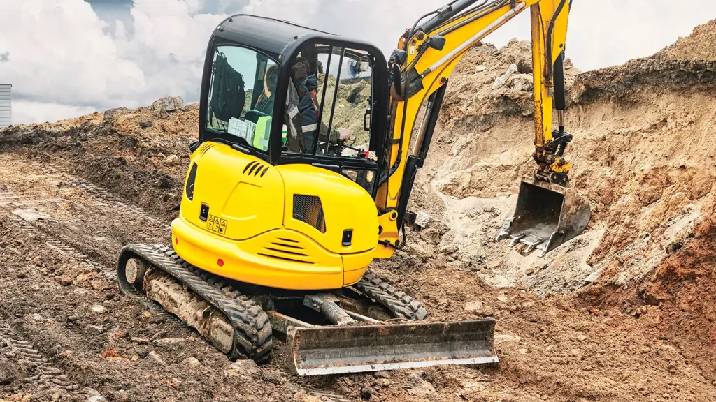 Yellow mini excavator with operator digging a trench in sandy soil